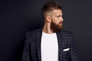 Handsome man with perfect hairdo and beard looking at side while standing against black background