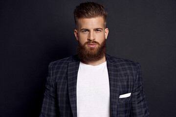 Handsome man with perfect hairdo and beard looking at camera while standing against black background