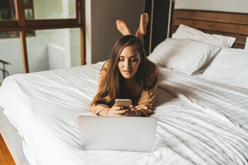 Obraz premium Woman lying on bed with laptop chatting in phone in bedroom