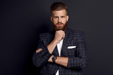 Young hipster holding hand on chin and looking at camera while standing against grey background