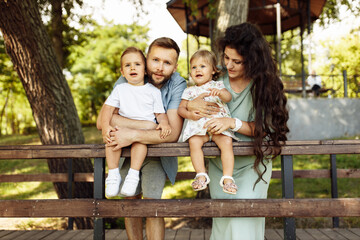 Fototapeta premium Lovely parents with little kids at the park, caring father hold in arms joyful son, cute daughter sitting in mothers hands, enjoy happy family moments, weekend outdoors concept