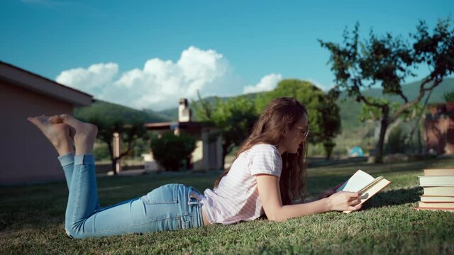 Distance education concept, young pretty female student studying at home, relaxing in the garden on the grass and reading interesting books, preparing online homework and doing information researches