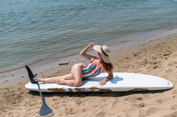 A woman in a swimsuit and a hat is posing lying on a sup board on the beach.