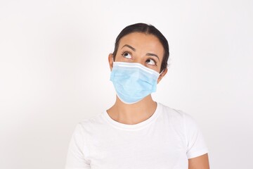 Young arab woman wearing medical mask standing over isolated white background, looking aside into empty space thoughtful