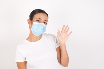 Young arab woman wearing medical mask standing over isolated white background Waiving saying hello...
