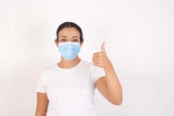 Young arab woman wearing medical mask standing over isolated white background doing happy thumbs up gesture with hand. Approving expression looking at the camera showing success.