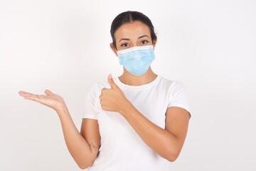 Young arab woman wearing medical mask standing over isolated white background Showing palm hand and doing ok gesture with thumbs up, smiling happy and cheerful.