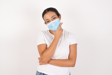 Young arab woman wearing medical mask standing over isolated white background looking confident at the camera smiling with crossed arms and hand raised on chin. Thinking positive.