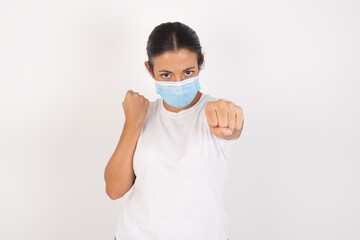 Young arab woman wearing medical mask standing over isolated white background Punching fist to fight, aggressive and angry attack, threat and violence