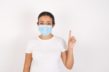 Young arab woman wearing medical mask standing over isolated white background showing and pointing up with finger number one while smiling confident and happy.