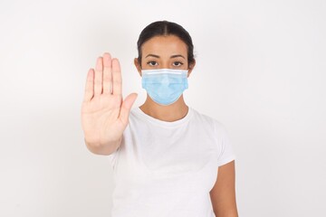 Young arab woman wearing medical mask standing over isolated white background doing stop gesture with palm of the hand. Warning expression with negative and serious gesture on the face