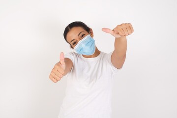 Young arab woman wearing medical mask standing over isolated white background approving doing positive gesture with hand, thumbs up smiling and happy for success. Winner gesture.