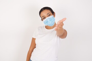 Young arab woman wearing medical mask standing over isolated white background smiling friendly offering handshake as greeting and welcoming. Successful business.