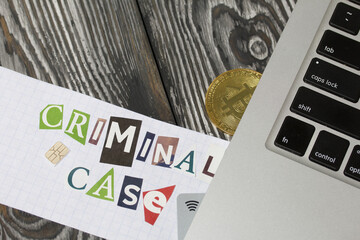 The phrase Criminal case made of letters cut from a magazine and pasted on a sheet of paper. Bitcoin and bank cards are sticking out of the laptop. On brushed pine boards painted black and white.