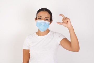Young arab woman wearing medical mask standing over isolated white background smiling and confident gesturing with hand doing small size sign with fingers looking and the camera. Measure concept