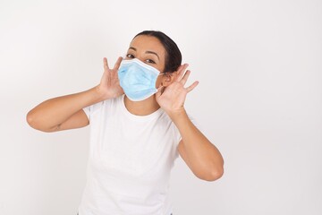 Young arab woman wearing medical mask standing over isolated white background Trying to hear both hands on ear gesture, curious for gossip. Hearing problem, deaf