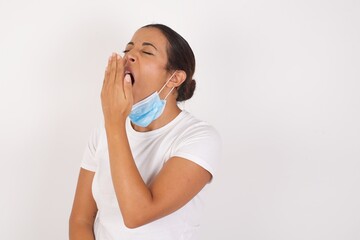 Young arab woman wearing medical mask standing over isolated white background being tired and yawning after spending all day at work.