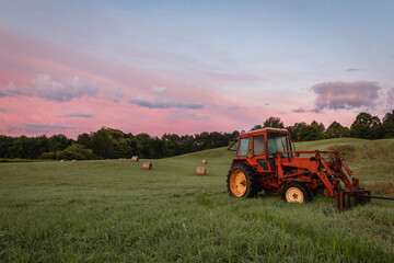 Obraz premium Red tractor and freshly rolled hay bales rest on rolling hill with dramatic cloudscape at sunrise
