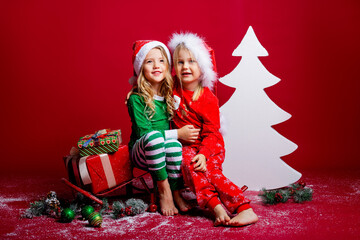 Two little girls on christmas hats on a red background sit near a white Christmas tree with gifts