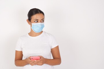 Young arab woman wearing medical mask standing over isolated white background hold telephone hands read good youth news look empty space advert