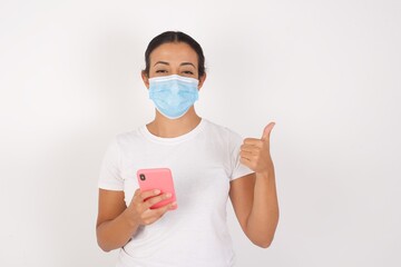 Young arab woman wearing medical mask standing over isolated white background using and texting with smartphone  happy with big smile doing ok sign, thumb up with fingers, excellent sign