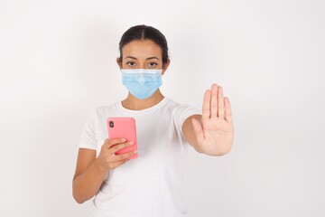 Young arab woman wearing medical mask standing over isolated white background  using and texting with smartphone with open hand doing stop sign with serious and confident expression, defense gesture