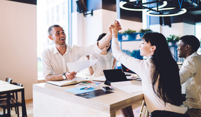 Positive multiracial colleagues giving high five while sitting at desk during meeting