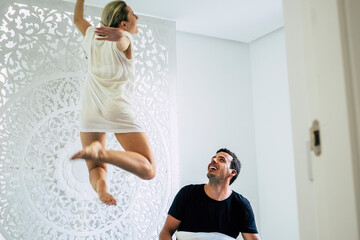 Fototapeta na wymiar Happy young caucasian couple enjoy and celebrate new home - happiness and joyful people concept with active and beautiful girl jumping on the bed and boyfriend looking with fun and a smile