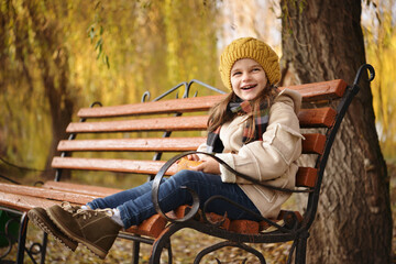 Cheerful preschooler little girl in coat and yellow hat with autumn leaves in the park. Child portrait. Cute kid girl 4-5 year old posing outdoors, has happy face. Walking in park. Childhood.