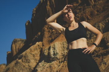 Fototapeta premium Tired exhausted young athletic woman runner wipes sweat from forehead, taking a rest, doing break, breathing hard after running hard at mountain landscape nature path in hot sunny day