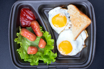 Fried eggs with sausages and croutons on a dark background. Scrambled eggs in a disposable plate close-up.