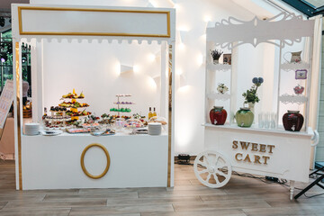 Full view of white designed sweet cart full of yummy baked desserts, fresh exotic fruits and non alcohol drinks. Children birthday party. Concept of healthy food on party, keto diet.