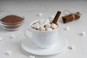Cup full of hot homemade cocoa drink usually prepared at cold winter season served on plate surrounded with marshmallows and cinnamon on white wooden background at kitchen. Horizontal orientation