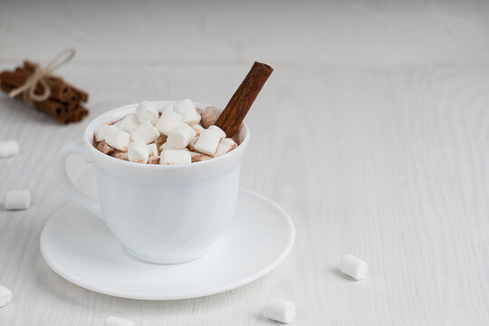 Cup full of hot homemade cacao drink usually prepared at cold winter and autumn seasons served on plate with marshmallows and cinnamon on white wooden background. Image with copy space, horizontal