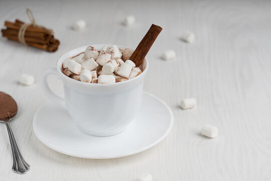Cup full of hot homemade cocoa drink usually prepared at cold winter and autumn days served on plate with marshmallows, cinnamon and spoon on white wooden background. Image with copy space, horizontal