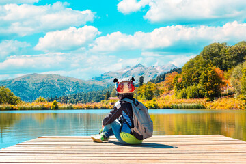 Autumn landscape. A person in a motorcycle jacket and helmet, sitting on the pier against the background of the lake and the autumn forest. Blue sky with clouds. Travel concept