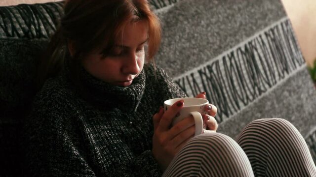A young caucasian woman is being treated for a cold with a decoction of medicinal tea. A sickly woman is warmed by hot tea while sitting on the couch. Selective focus.