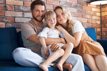 Fototapeta premium portrait of adorable young family with child girl, they sit on sofa together in living room, have rest, love