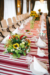 Front view of restaurant table serving in cozy rustic style on authentic table covering. Preparing for traditional wedding celebration. Table floral centerpieces with fresh sunflowers and greenery.