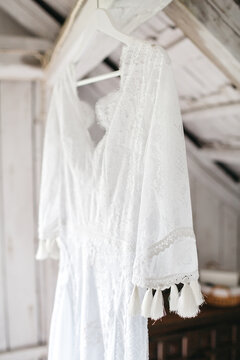 Front view of hanging white dress in boho style in lightroom. Charming clothing for fiance with lace and embroidery. Sewing individual outfit for formal occasion. Concept of pretty bridal fashion.