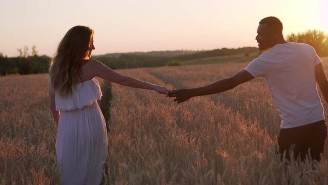 Young pregnant caucasian woman and her african husband walk holding hands in a wheat field at sunset. View from the back.