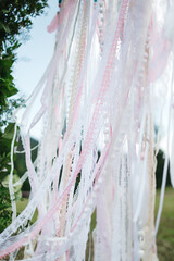 Close up of tender white and pale pink ribbons and feathers on ceremonial wedding arch on green backyard waving on wind. Dream catcher at bridal zone design. Concept of boho style and tenderness.
