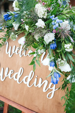 Close up of welcome wedding wooden tablet decorated with blue, white flowers and greenery on celebration. Beautiful floral composition on unique bridal furniture. Concept of wedding preparation detail