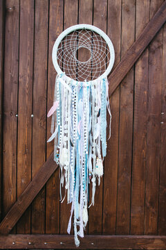 Front view of beautiful dream catcher made of pale ribbons, feathers and beads hanging on wooden wall. Original handmade decor details for wedding celebration in boho style. Concept of unique party.