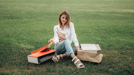 Obraz premium Portrait of european lady hold hand colorful bags woman after shopping in white shirt jeans retail store near mall on the street sitting green grass park lawn sales black friday season blur background
