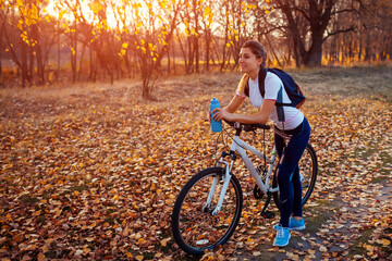Riding bicycle in autumn forest. Young woman having rest after workout on bike holding water bottle. Healthy lifestyle