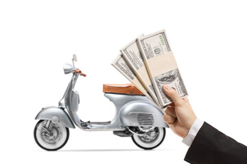 Hand of a young man in a suit holding stacks of us dollar money and a silver vintage scooter in the back