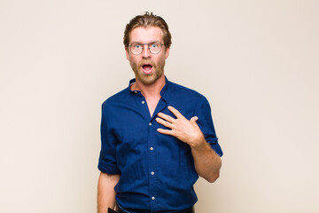 blonde adult caucasic man feeling shocked, astonished and surprised, with hand on chest and open...