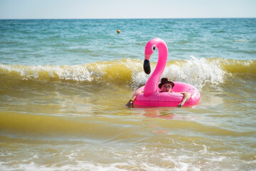 A girl in a swimsuit swims in the sea on an inflatable pink flamingo circle. The waves in the sea.