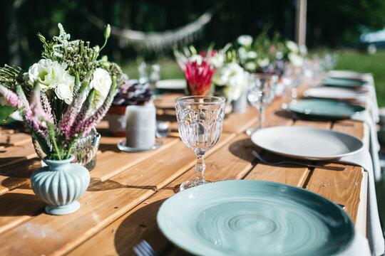 Close up of blue plates on wooden table, old fashioned goblets, tiny floral bouquets on backyard wedding celebration. Jubilee party outdoors. Decoration on table serving. Concept of family traditions.